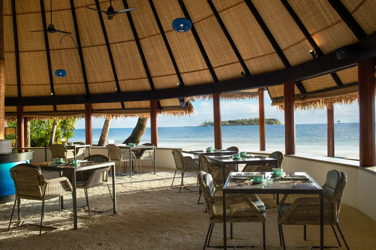 Riveli’s dining room under its thatched roof, tables laid and the turquoise sea beyond the open edge.