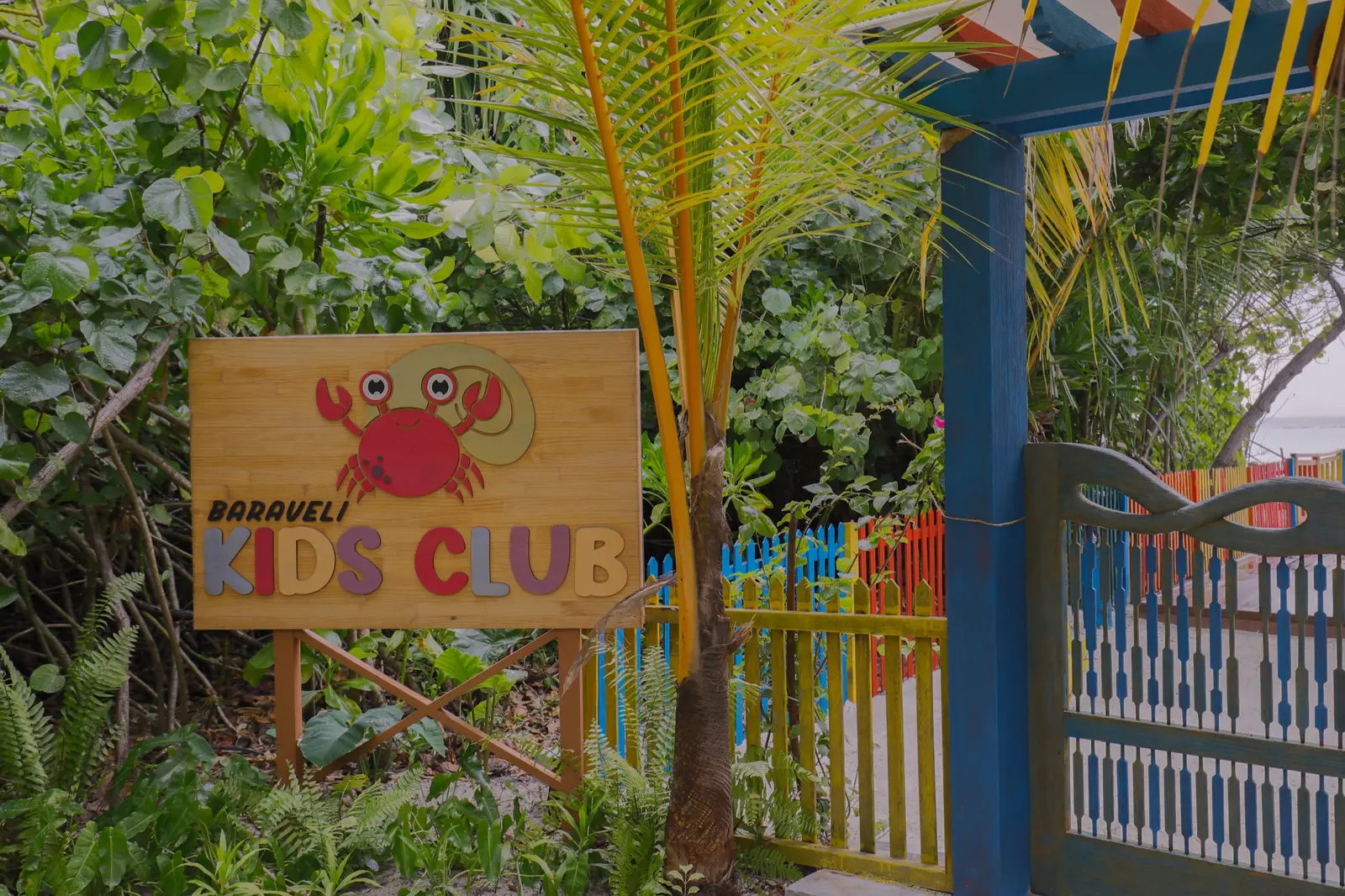 The Baraveli Kids Club sign and its painted gate among the plants.