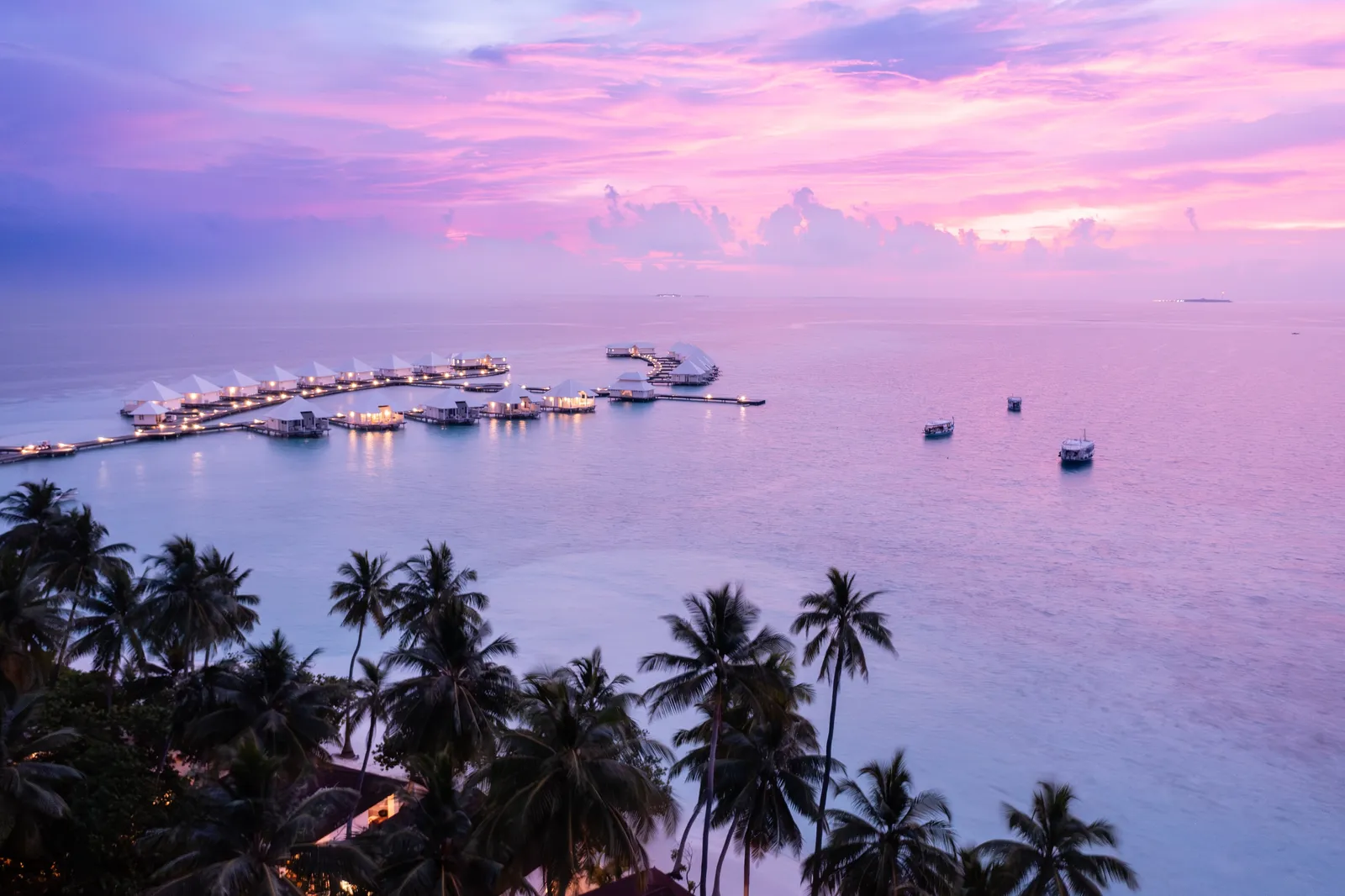 The water villas lit at dusk under a pink sky, with palms in the foreground.