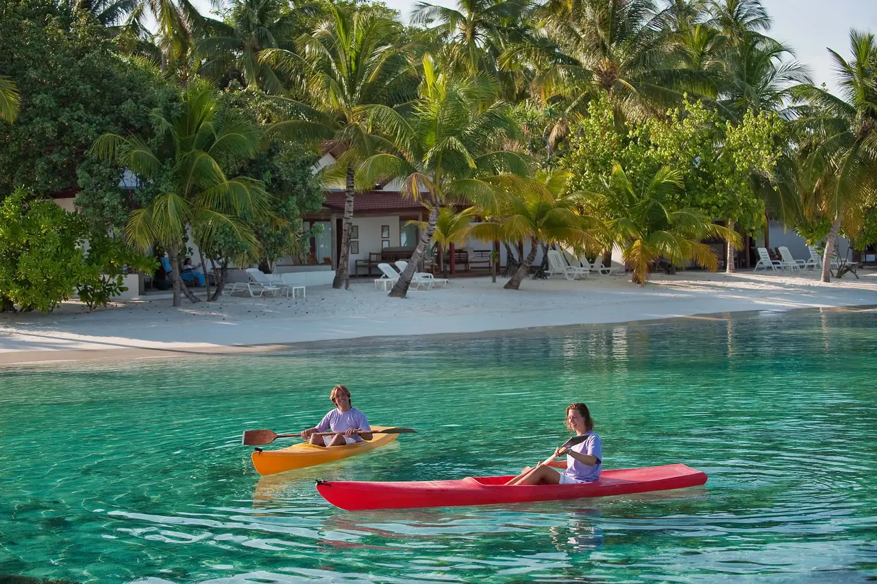 2 guests kayaking in the emerald lagoon, palms and villas on the shore.
