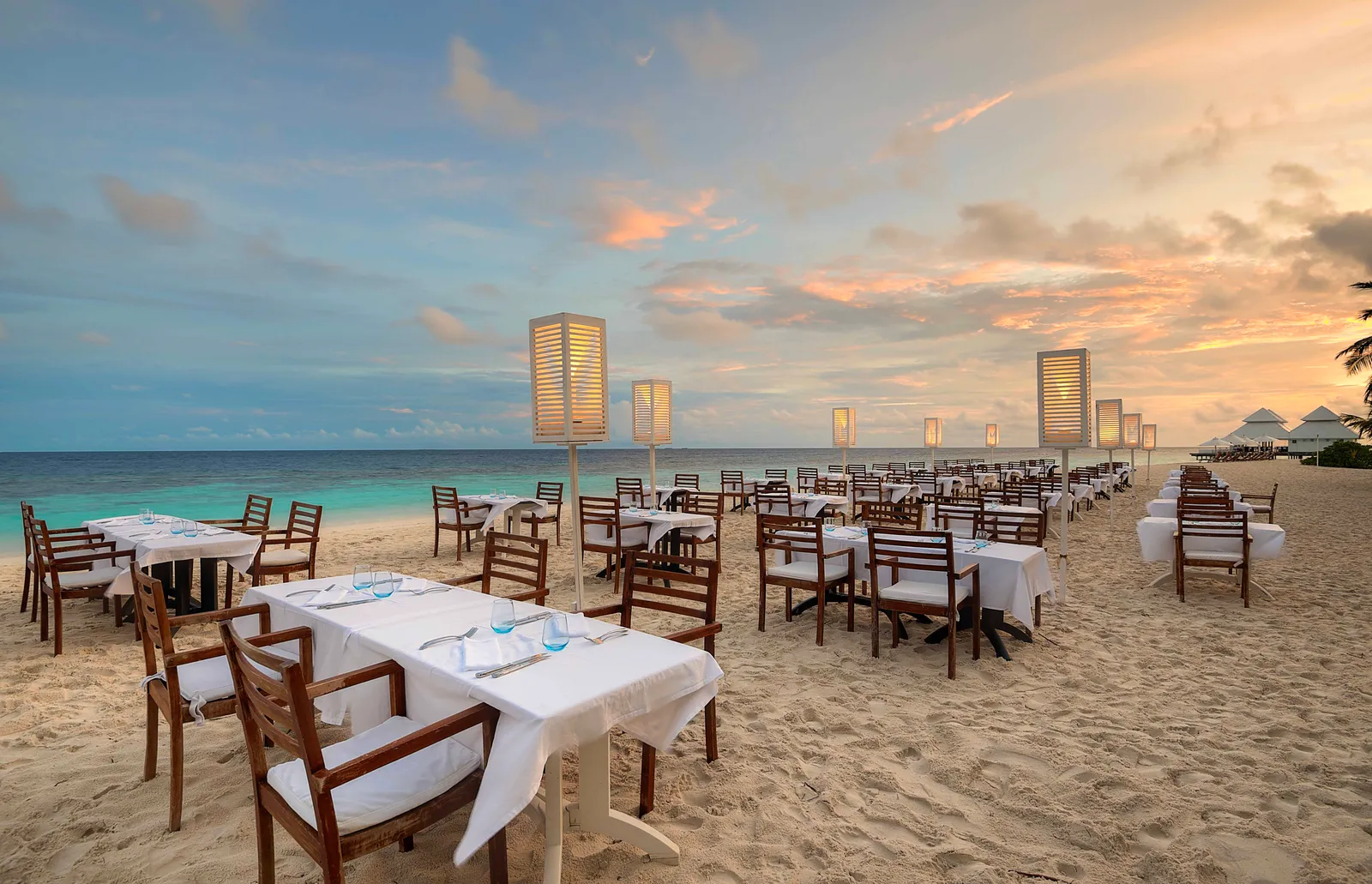 A Maldivian dinner on the beach at sunset, tables laid on the sand and lanterns.