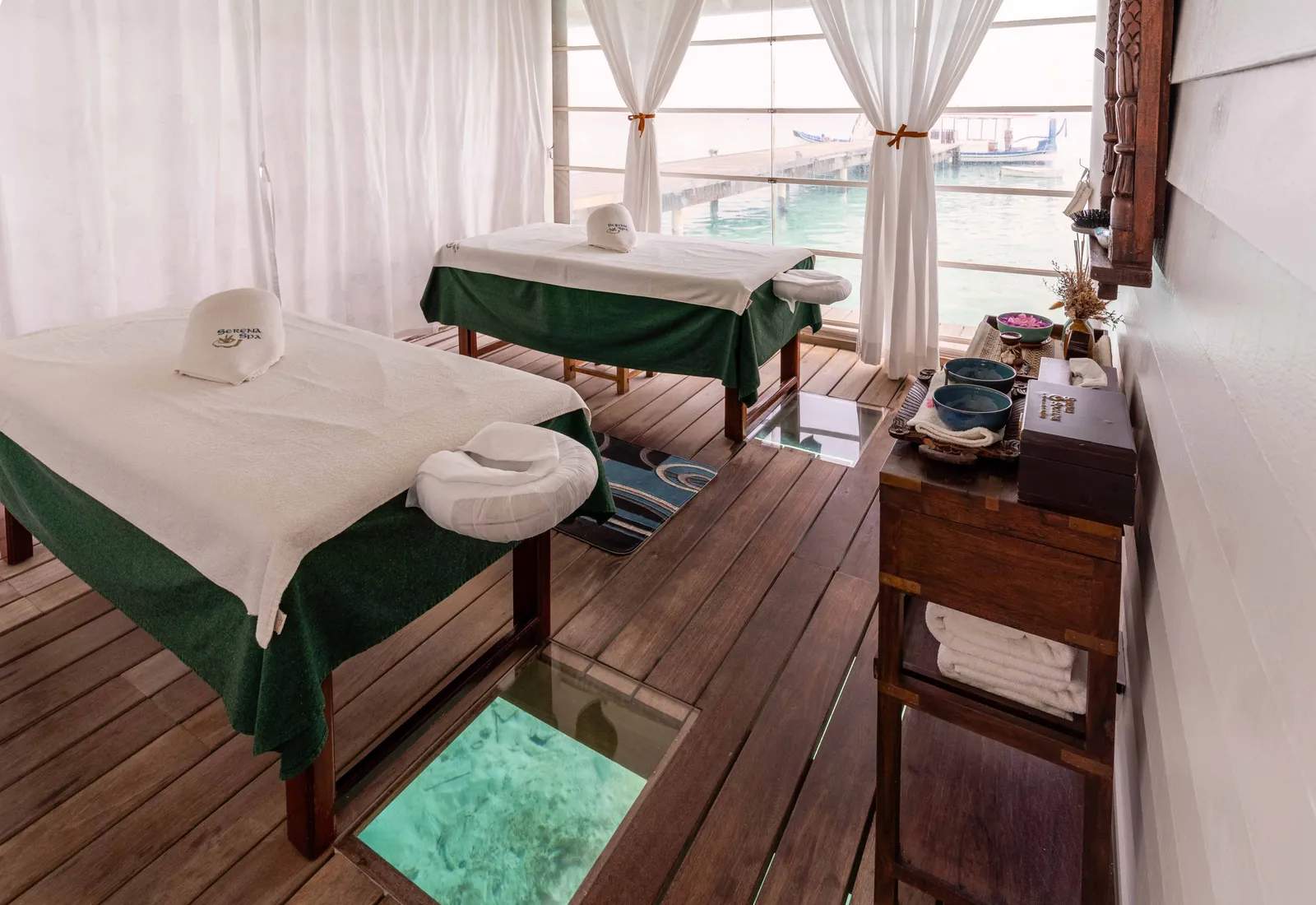 A Serena Spa room with 2 massage couches and a wooden floor with a glass panel over the lagoon.