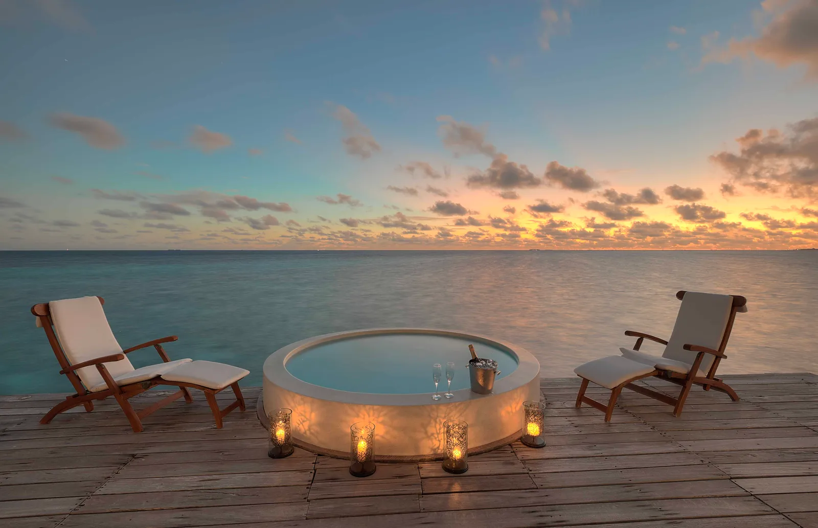 A round jacuzzi on a deck over the water at sunset, between 2 loungers and candles.