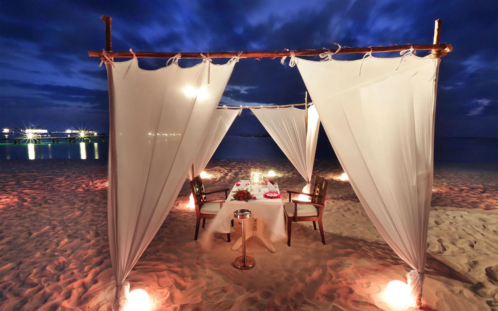 A table for 2 on the beach at night, between white drapes and lanterns.