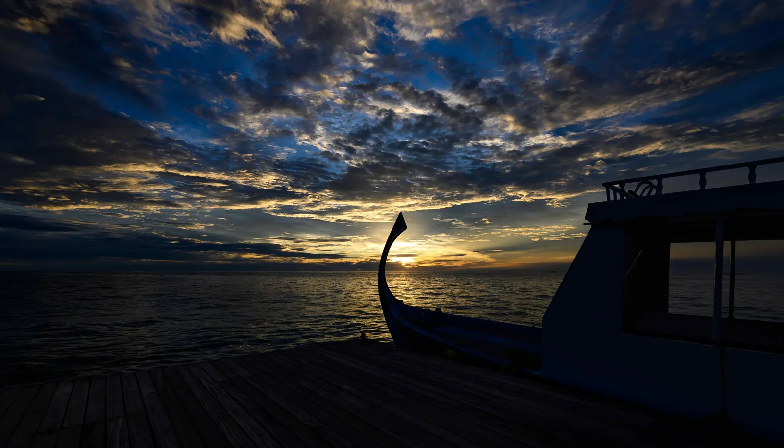 A dhoni, the Maldivian boat, silhouetted at sunset under a clouded sky.
