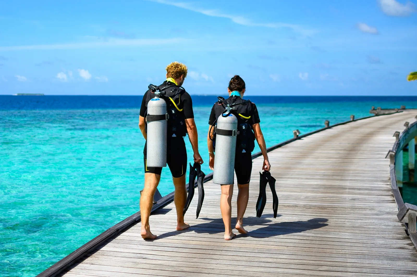 2 divers with their tanks walking along the jetty towards the sea.