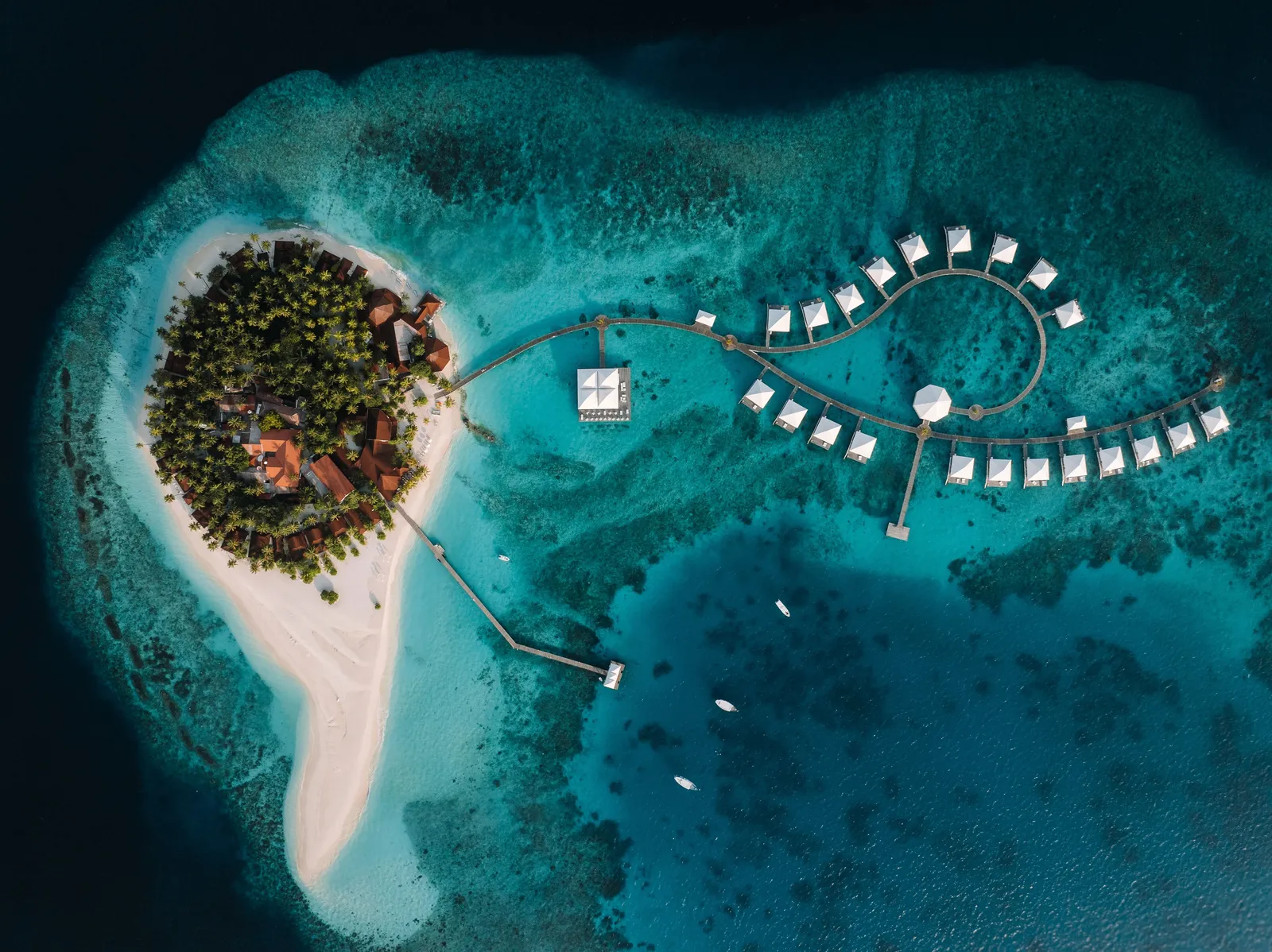 Thudufushi from above: the green island with its sand spit, the curving jetty of the water villas and the reef around it.