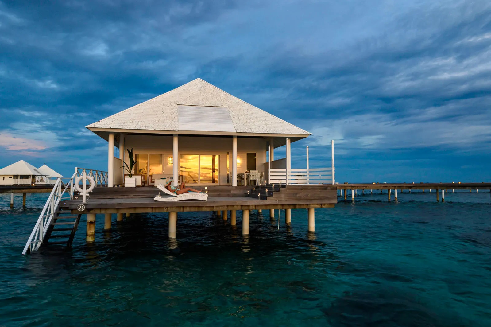 A water villa at dusk, lit from within, with its ladder down into the lagoon.