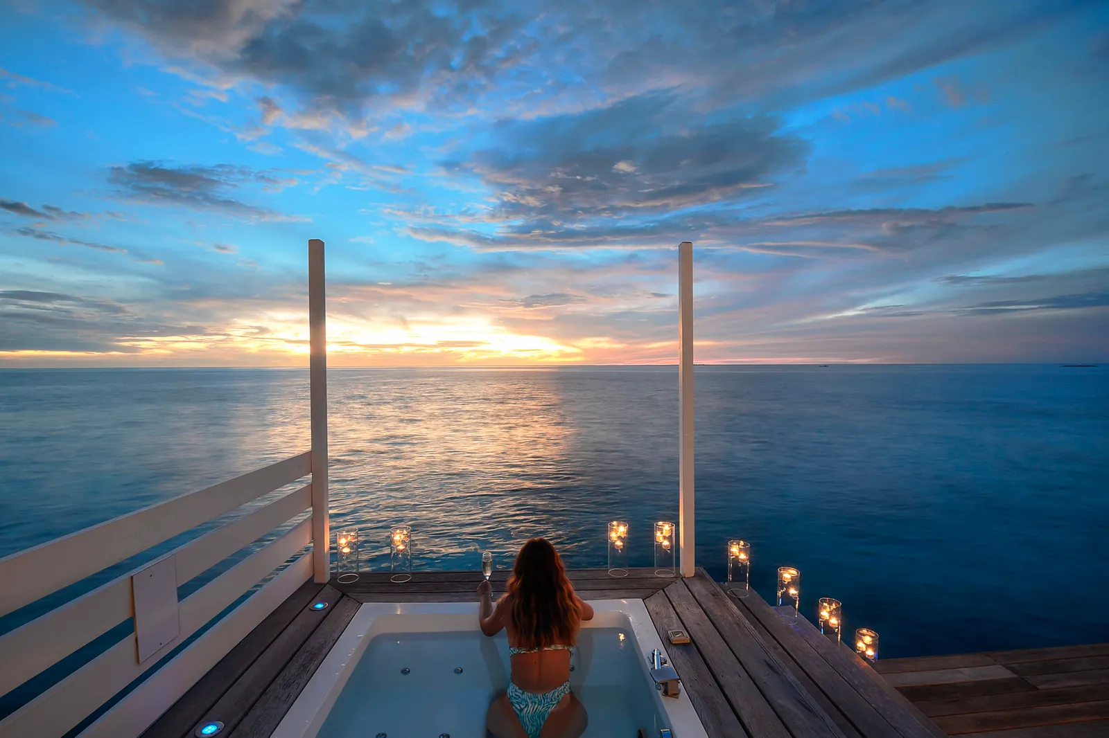 A guest in the jacuzzi on a Jacuzzi Water Villa’s deck at sunset, among lit candles.