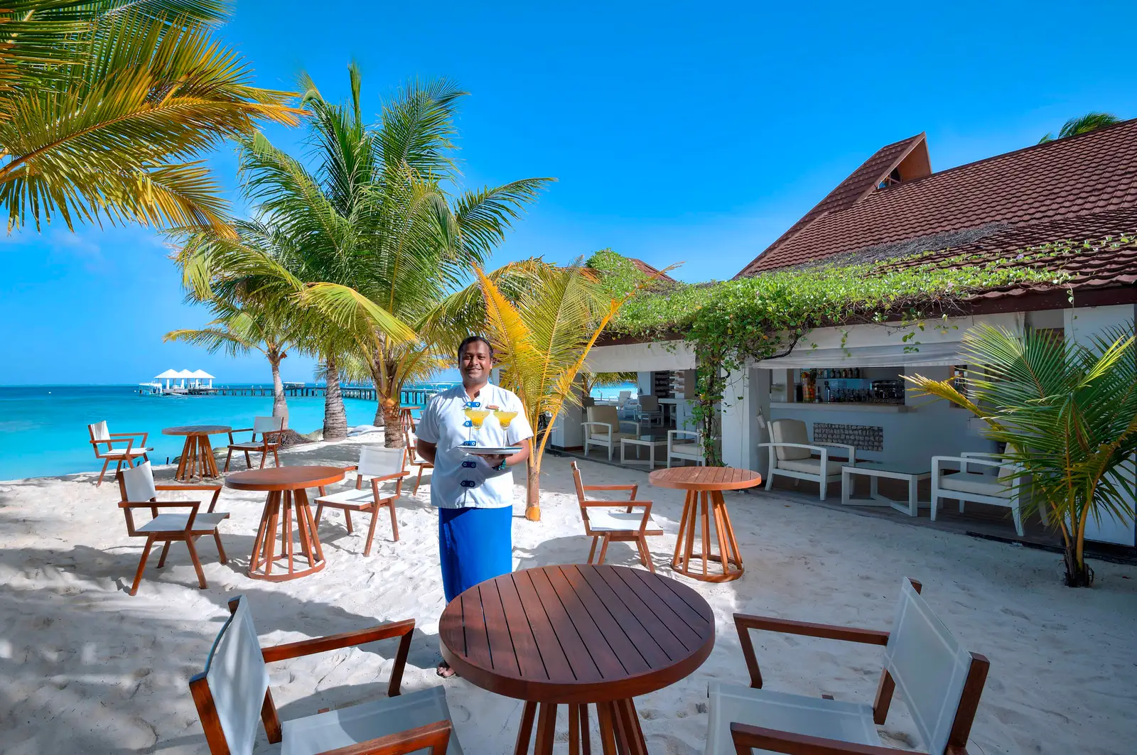 Veli Beach Bar on the sand, with its round tables, the palms and the lagoon.