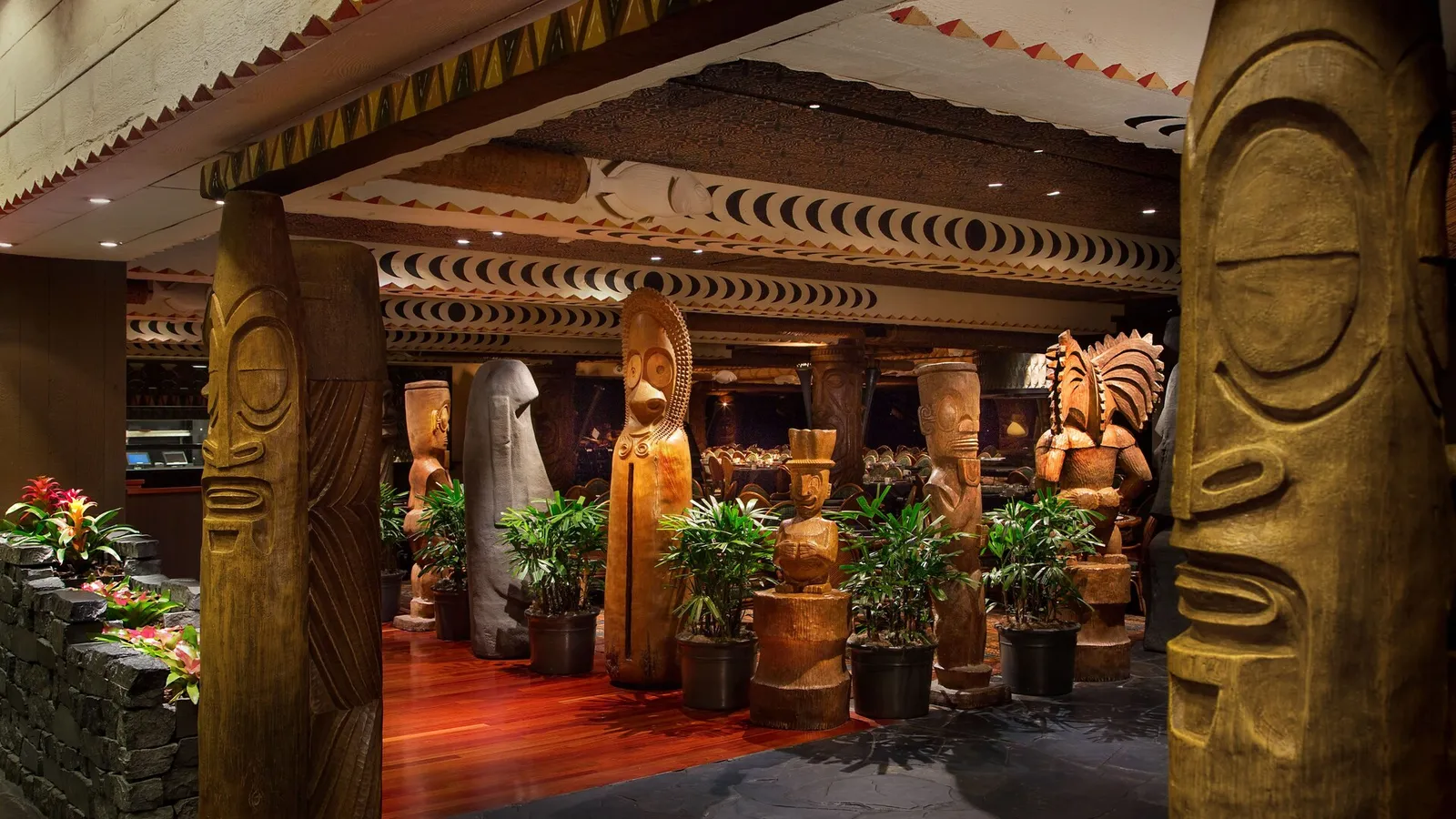 ʻOhana’s entrance between carved wooden tiki figures, plants and a red floor.