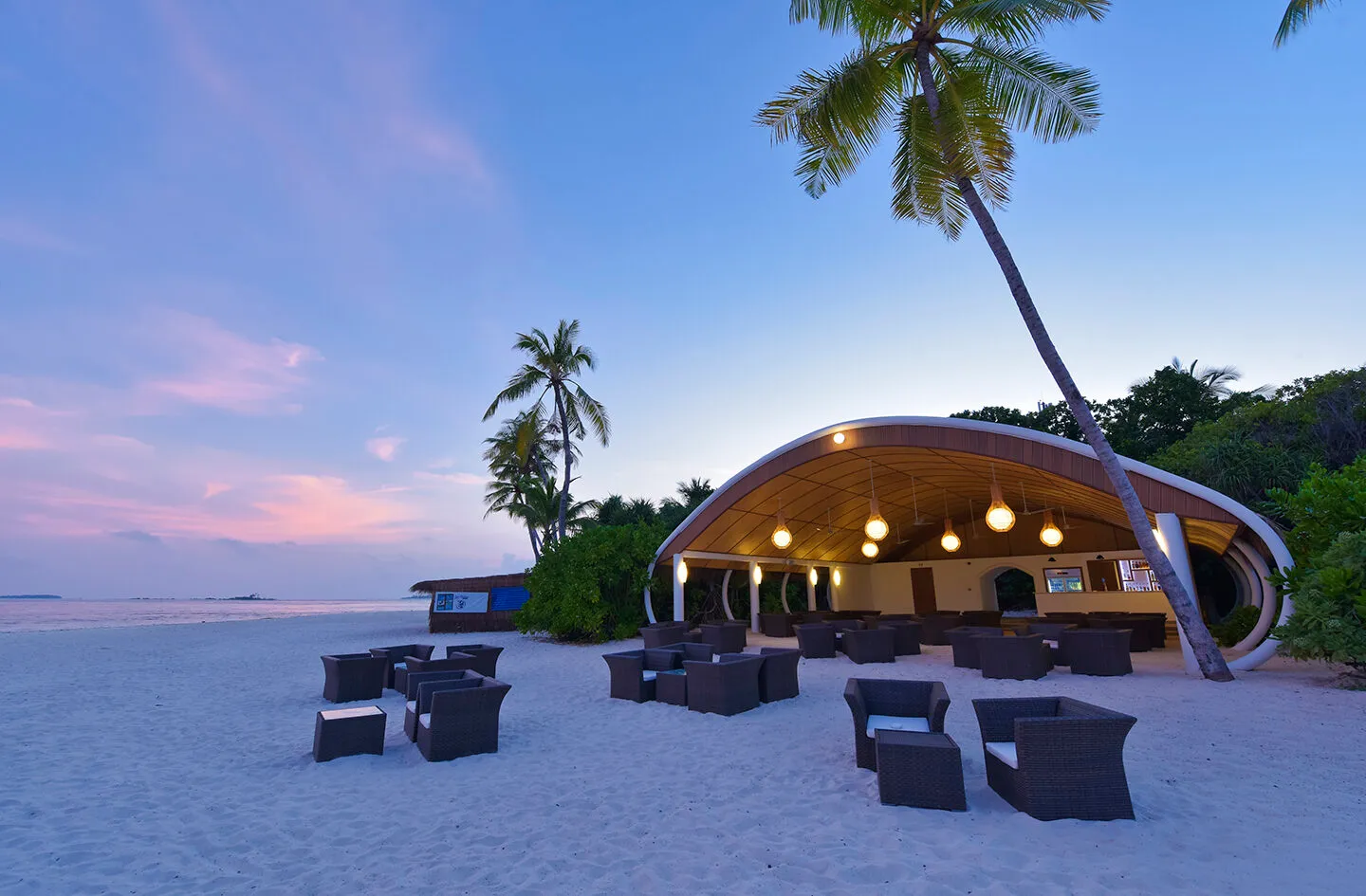 The Beach Bar on the sand at sunset, its curved roof lit, wicker armchairs and palms against a pink sky.