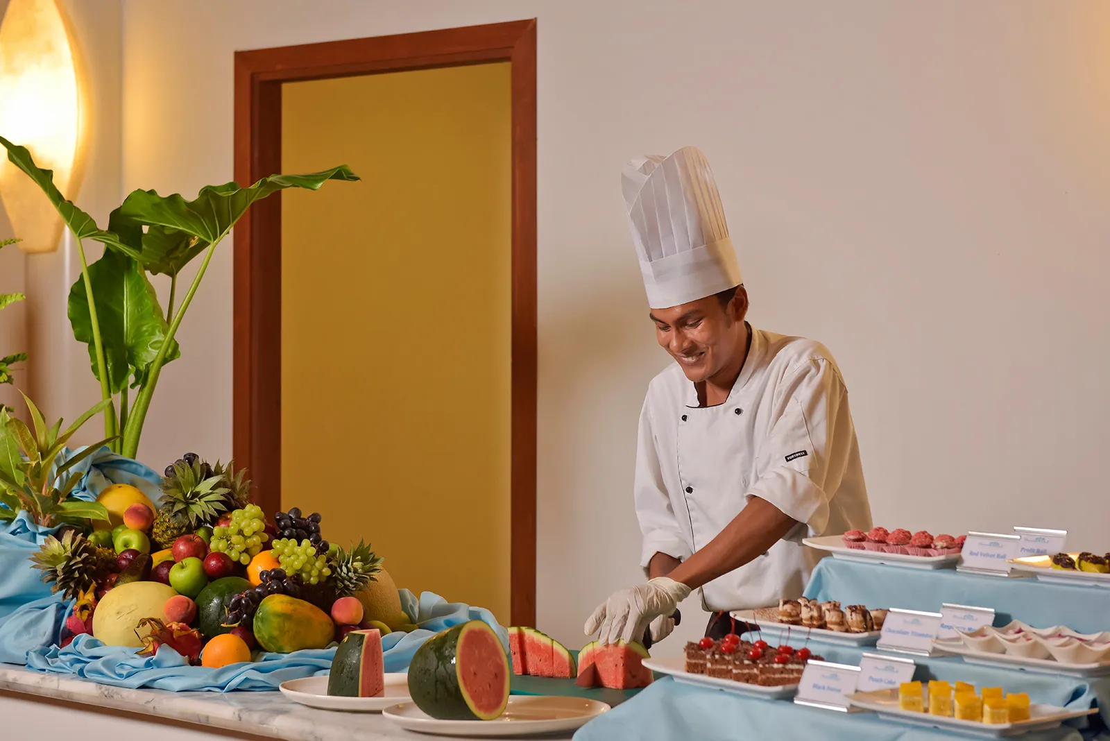 A chef carves fruit at the buffet, beside a basket of tropical fruit and trays of desserts.