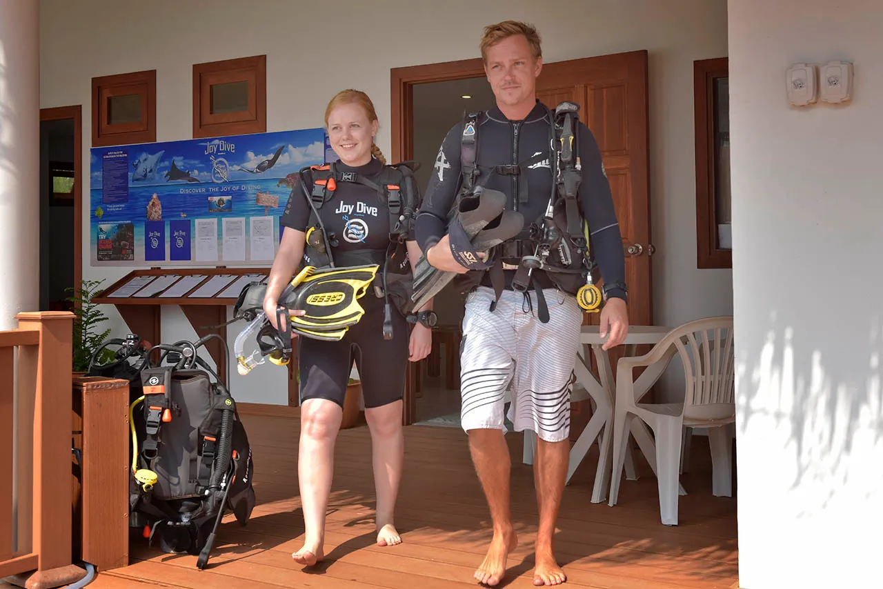 2 divers in wetsuits, carrying tanks and fins, walk out of the Joy Dive centre.