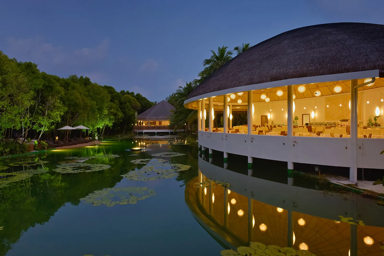 A round thatched pavilion with its tables laid, lit above the lake at dusk and mirrored in the water among the lilies.