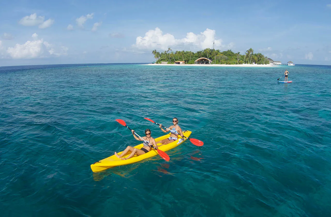 2 guests in a yellow kayak on the turquoise lagoon, someone on a paddleboard further off and the island behind.