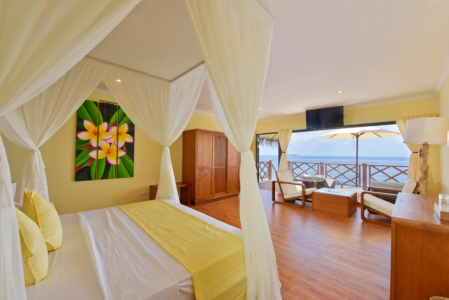 A water bungalow bedroom: the four-poster bed with white drapes, a painting of flowers and the deck over the sea.