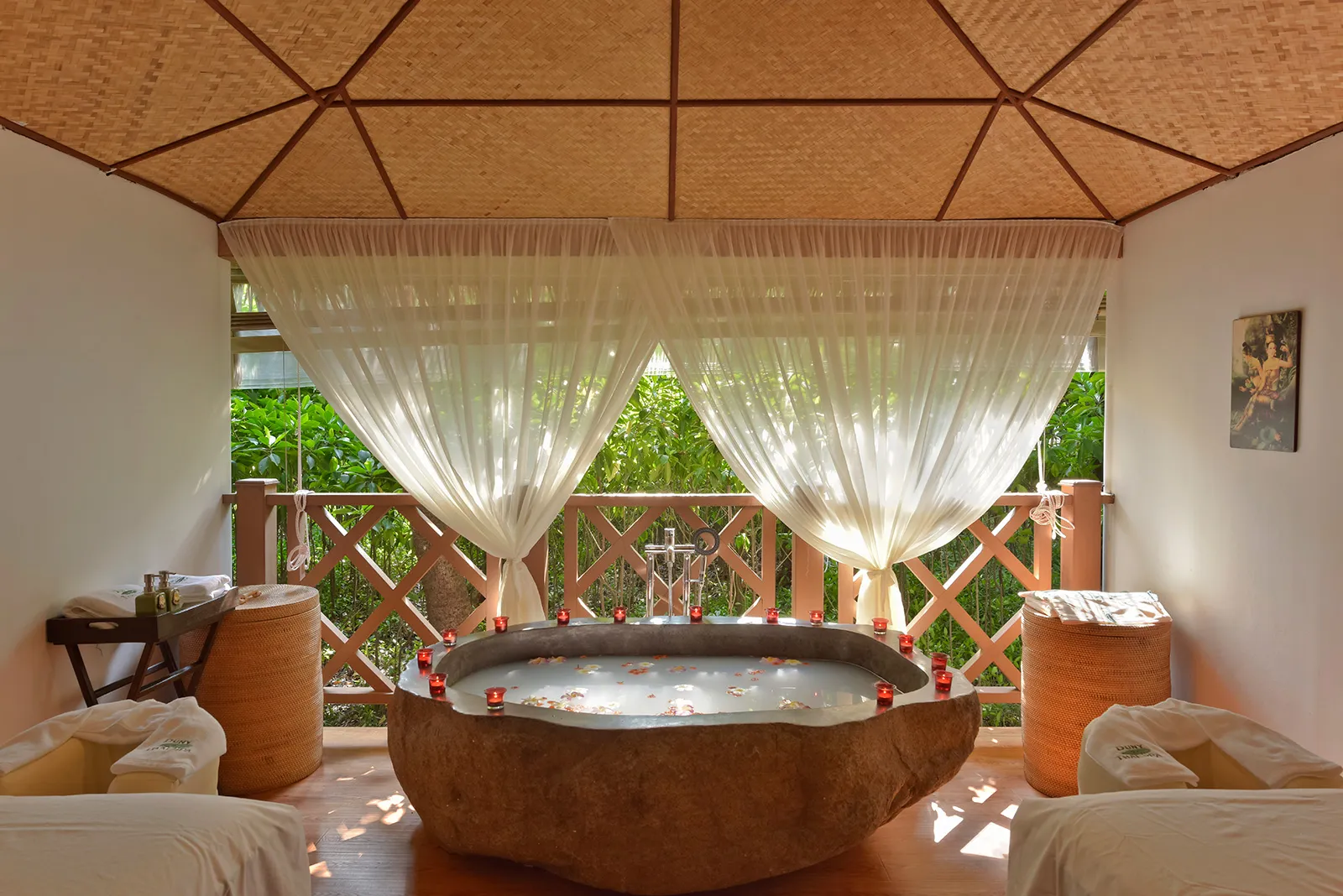 A spa room with a stone tub full of petals between 2 massage beds and the curtains open onto the green.