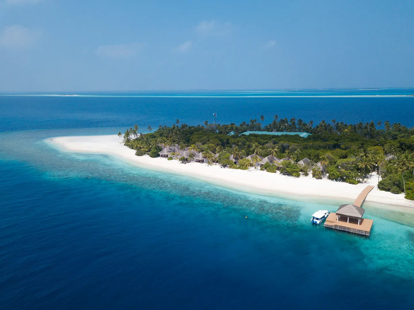 The long side of the island from above: the white beach, the bungalows among the palms, the arrival jetty with a speedboat and the reef in the shallows.
