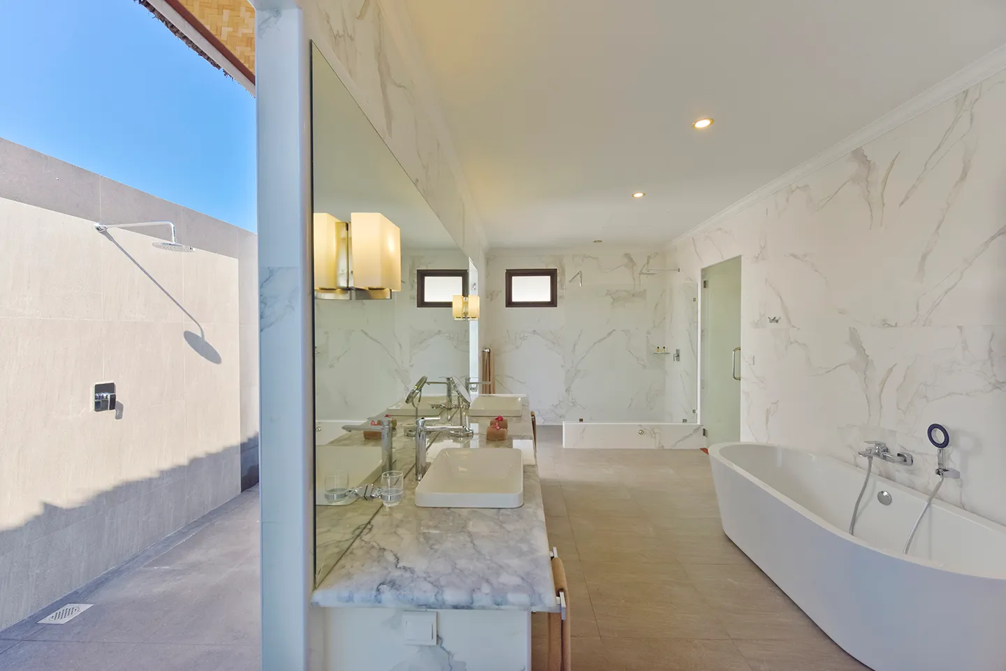 A water bungalow bathroom in pale marble, with a freestanding tub, 2 basins and the door to the outdoor shower.