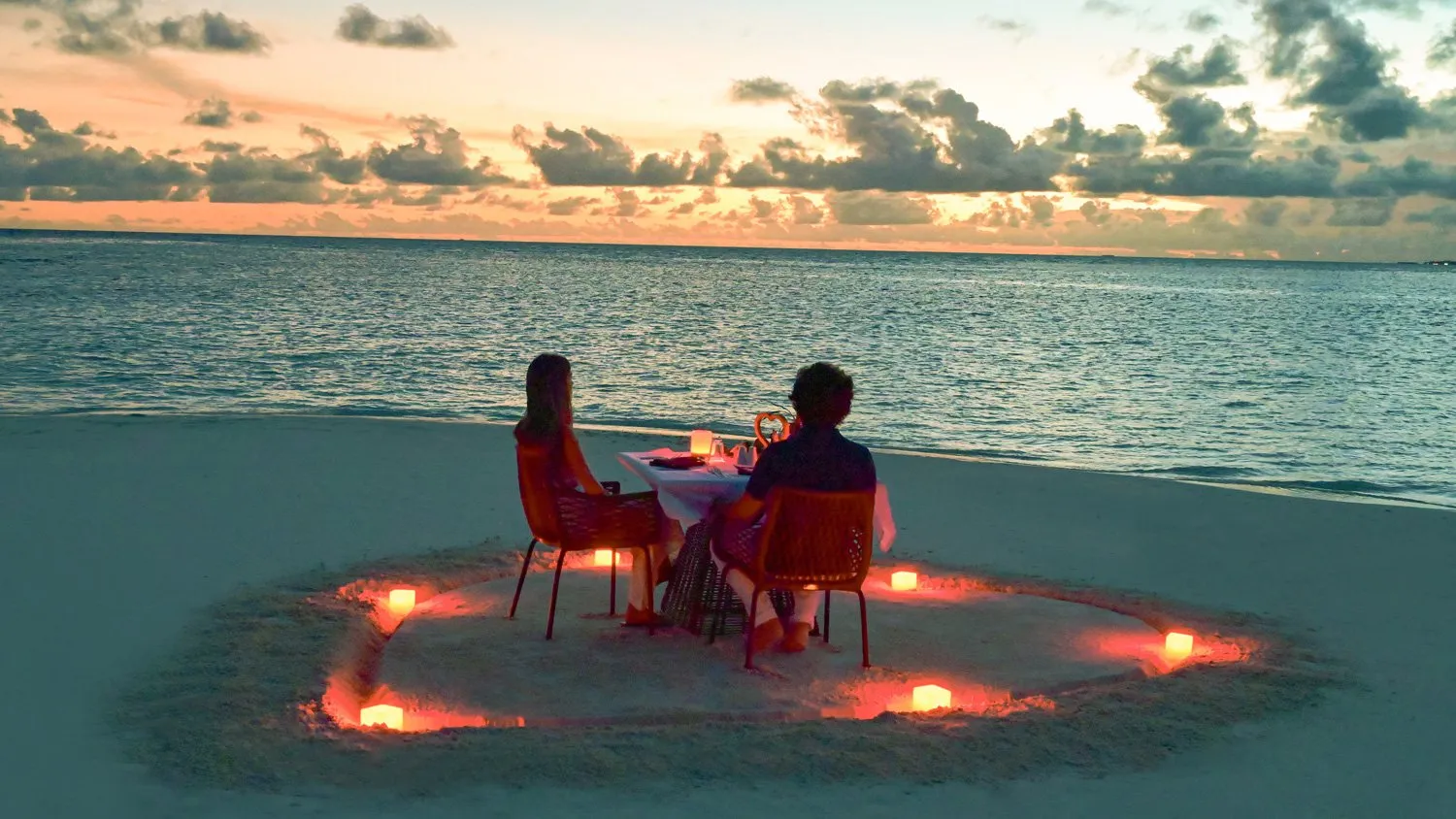 A couple dining on the beach at sunset, inside a ring of candles on the sand.