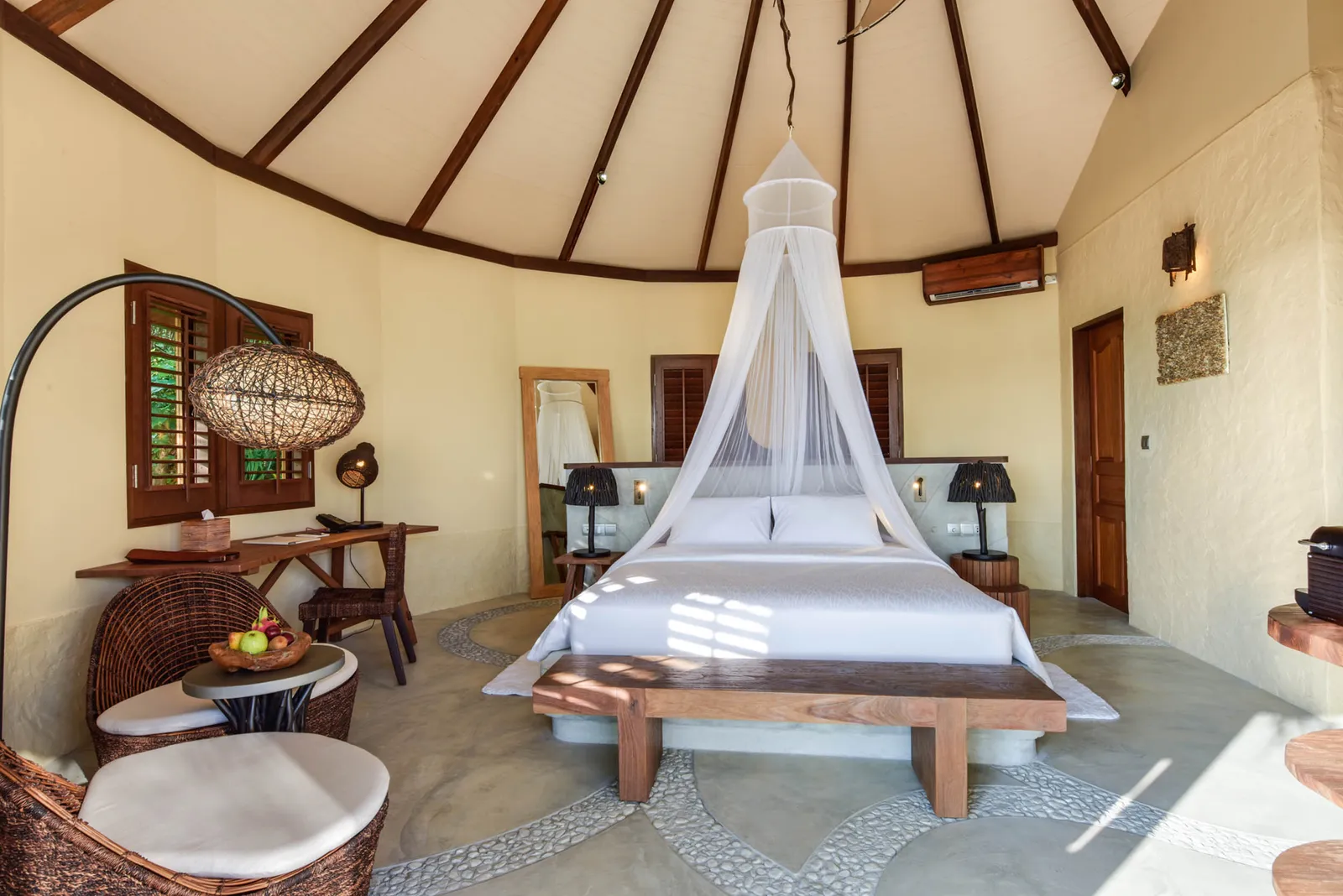 Inside a round Beach Villa: the bed under its net canopy beneath the timber ceiling, with carved furniture.