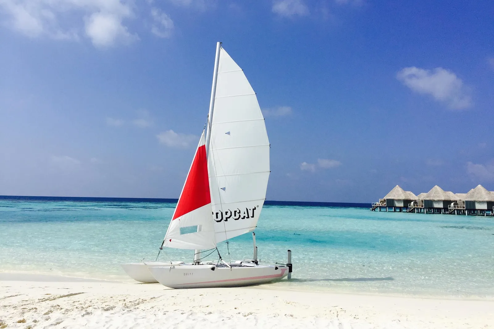 A catamaran with a red and white sail drawn up on the beach, the water villas in the background.