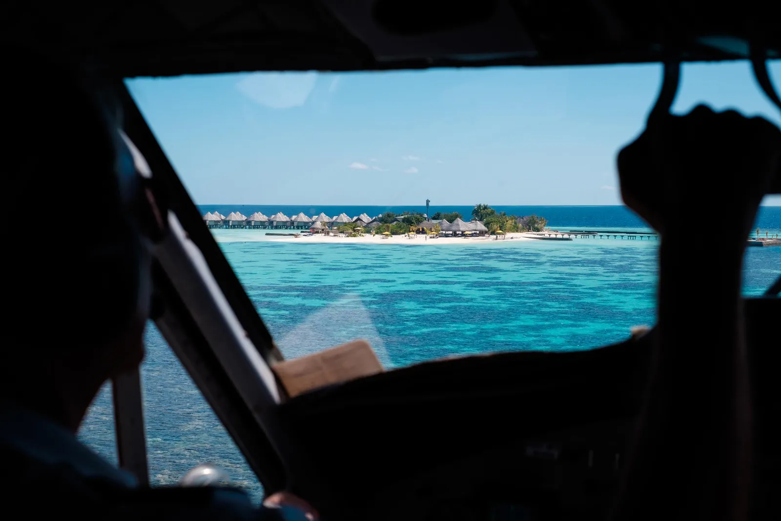 The island seen from the seaplane cockpit, between the pilot’s silhouette and his arm, the water villas on the horizon.