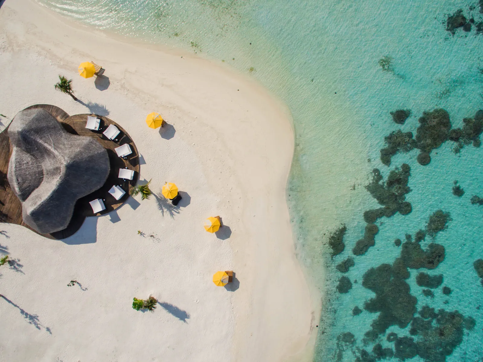 The beach from above, with yellow parasols, a thatched roof ringed by tables and the dark patches of reef a few metres off the shore.
