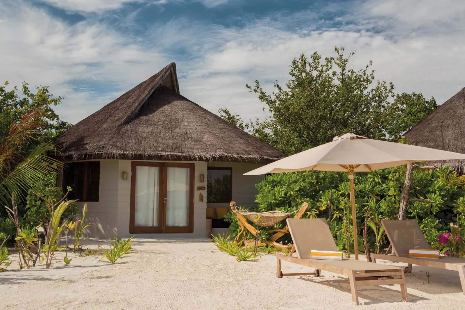 A Beach Villa under its thatched roof among the palms, loungers and a parasol on the sand in front.
