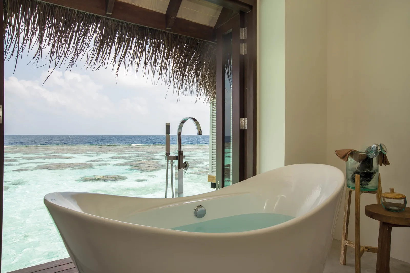 A Water Villa’s freestanding bath facing the open door, with the lagoon and the horizon beyond.