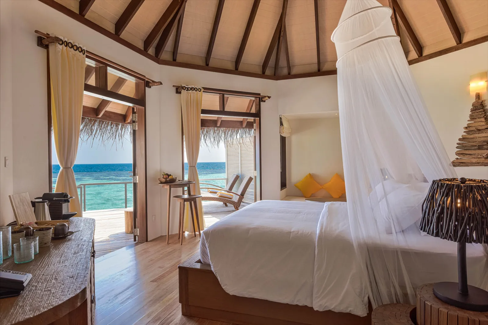 A Water Villa bedroom: the bed under its net canopy, yellow cushions and the doors open onto the deck and the sea.