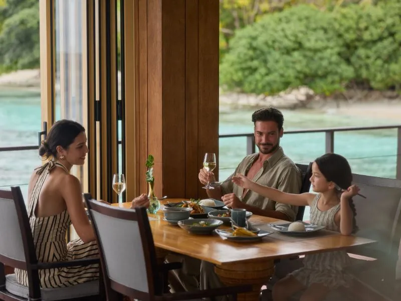 A family at table in Benjarong, with Thai dishes, a glass of wine and the lagoon behind the windows.