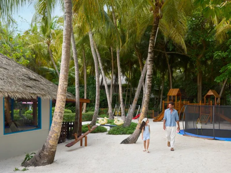 The kids’ club garden under the palms, with swings and slides and a father walking with his daughter.
