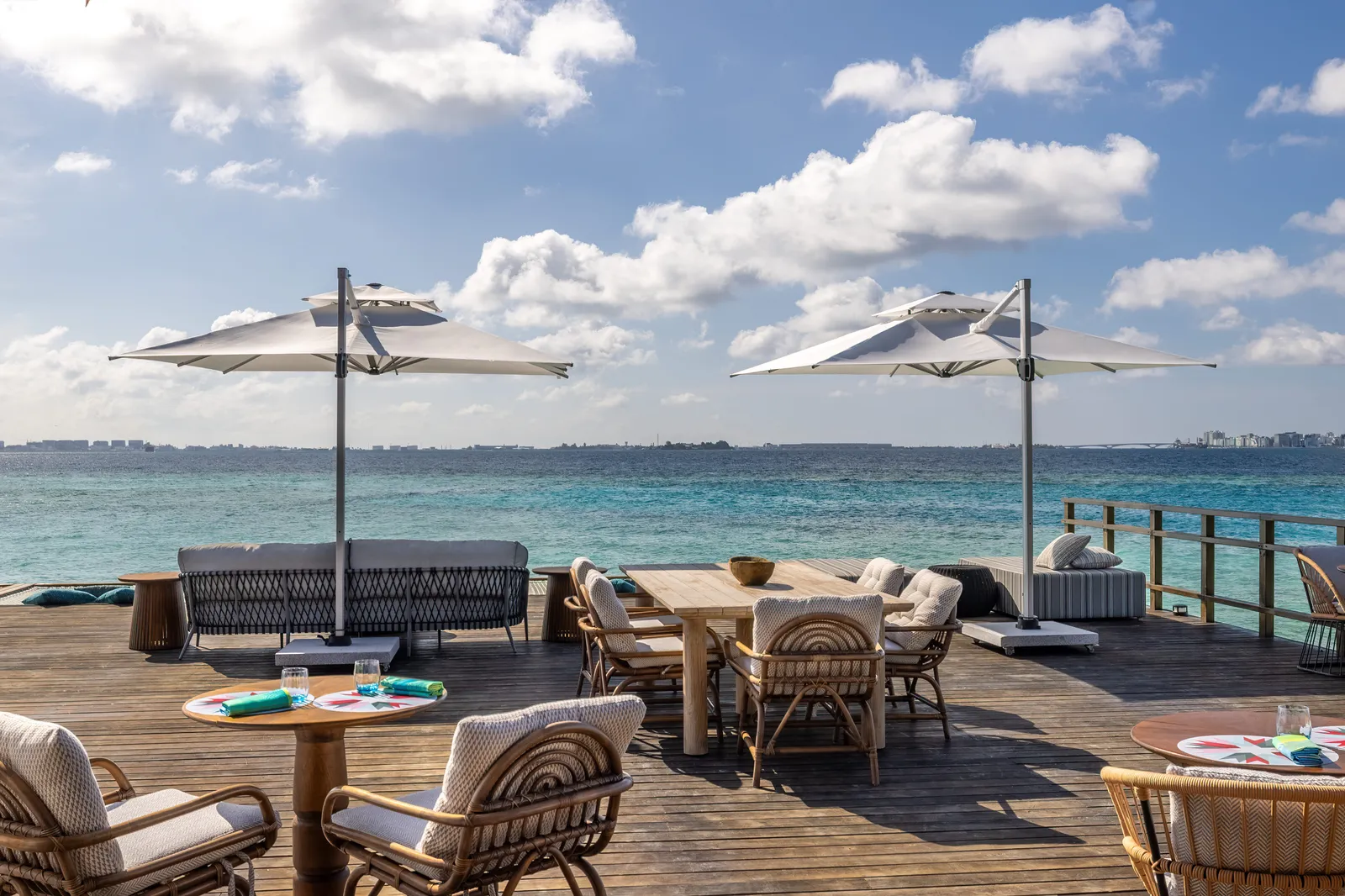 Cocoon’s deck over the sea, with tables, wicker armchairs, parasols and a line of buildings on the horizon.