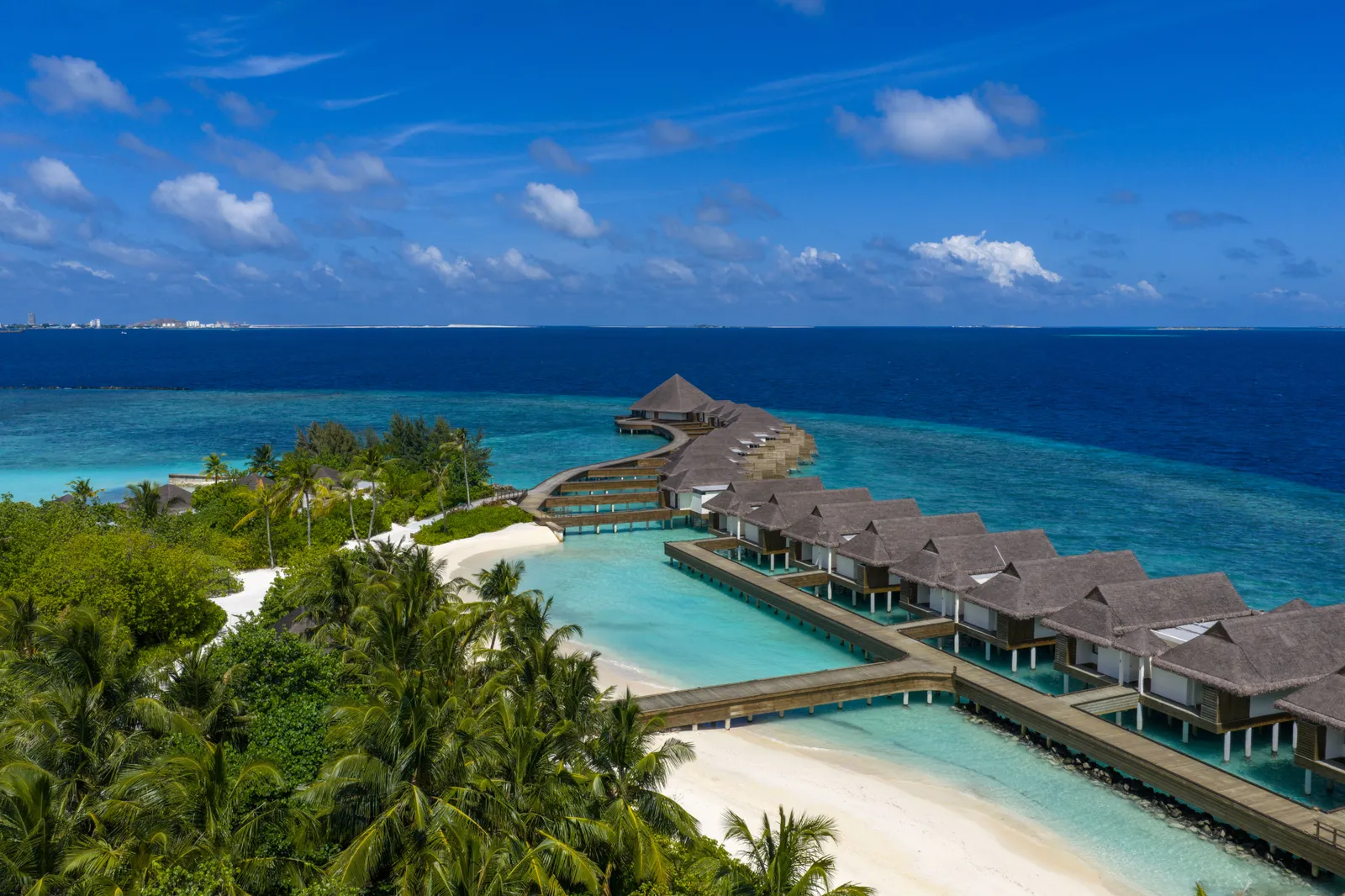 The island from above: the beach, the lagoon and the water villas, with open ocean and distant buildings on the left of the horizon.