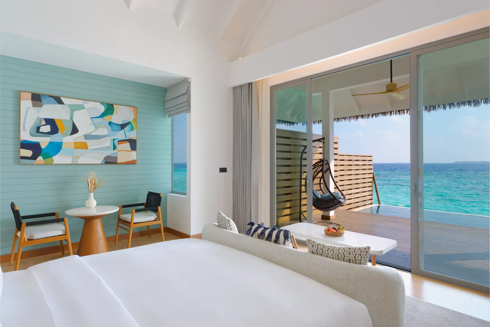 An Overwater Pool Villa: the bed, a small table, a hanging chair and glass doors open on to the deck above the ocean.