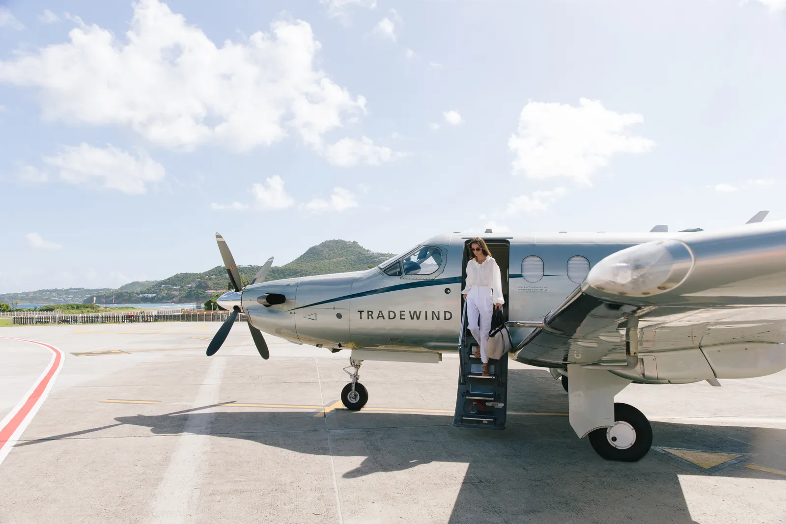 A small propeller aircraft on the airport apron, a passenger in white on its steps and the hills beyond.