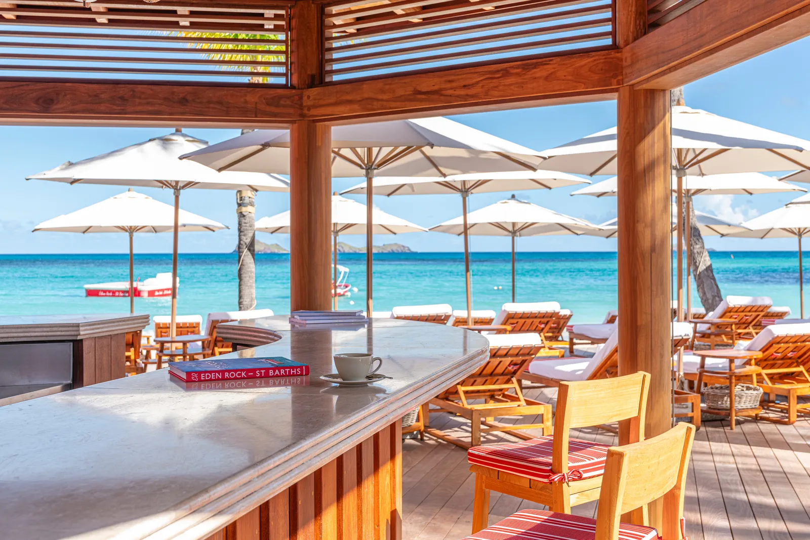 The Beach Bar’s counter from inside: timber posts, white parasols, loungers and the sea.