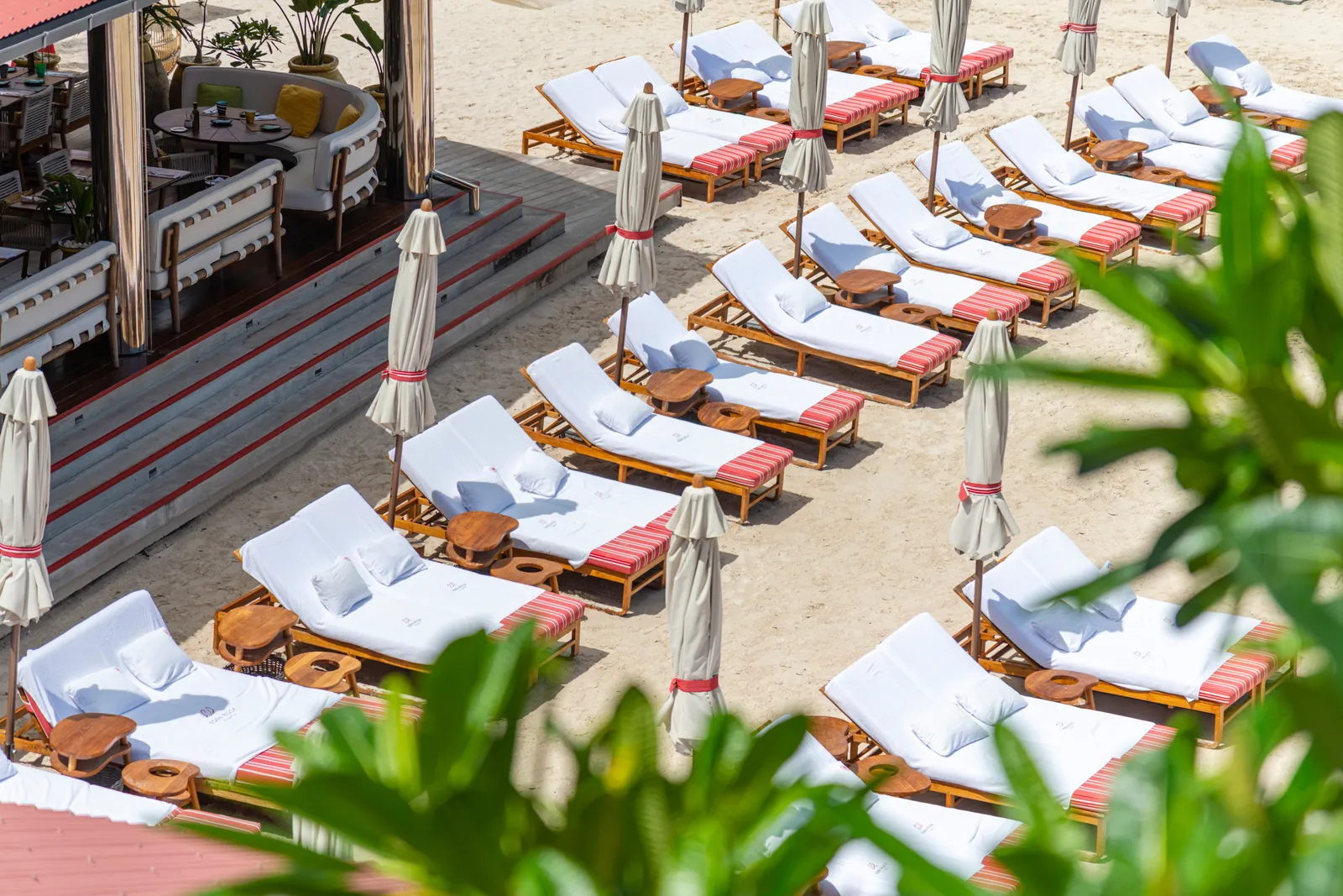 The beach from above: rows of loungers with white cushions and red-striped towels, parasols furled on the sand.