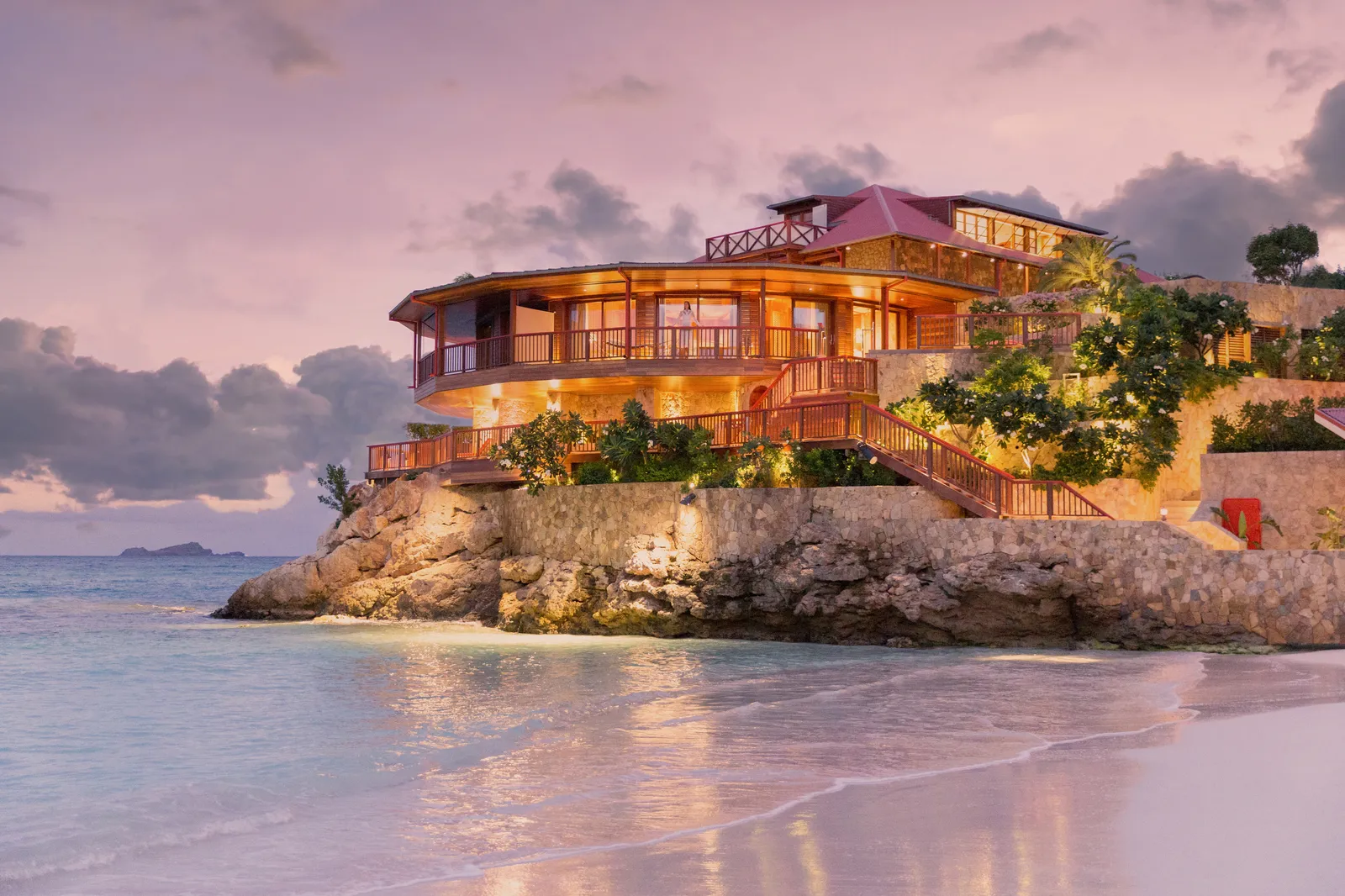 The hotel lit up on its rock at dusk, seen from the water’s edge under a pink and violet sky.