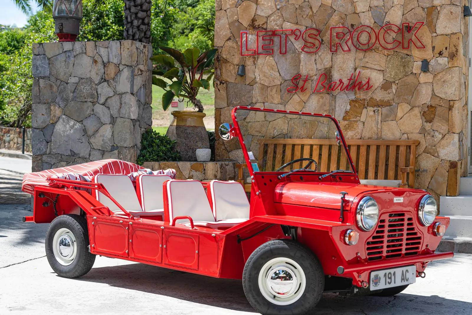 An open red Mini Moke parked by a stone wall with the neon sign «Let’s Rock St Barths».