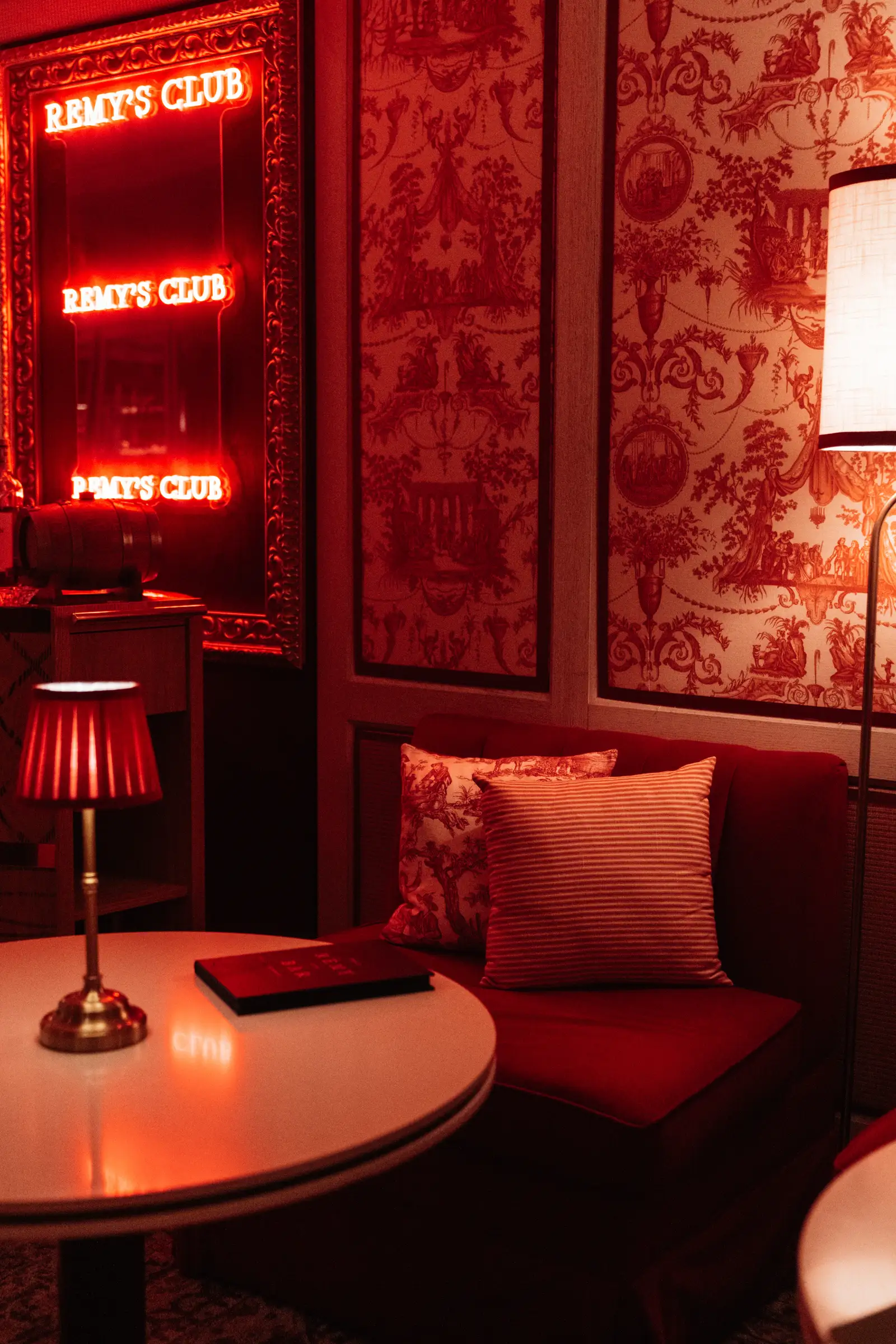 A corner of Rémy’s Club: red fabric walls, neon signs, a round table with a lamp and a banquette.