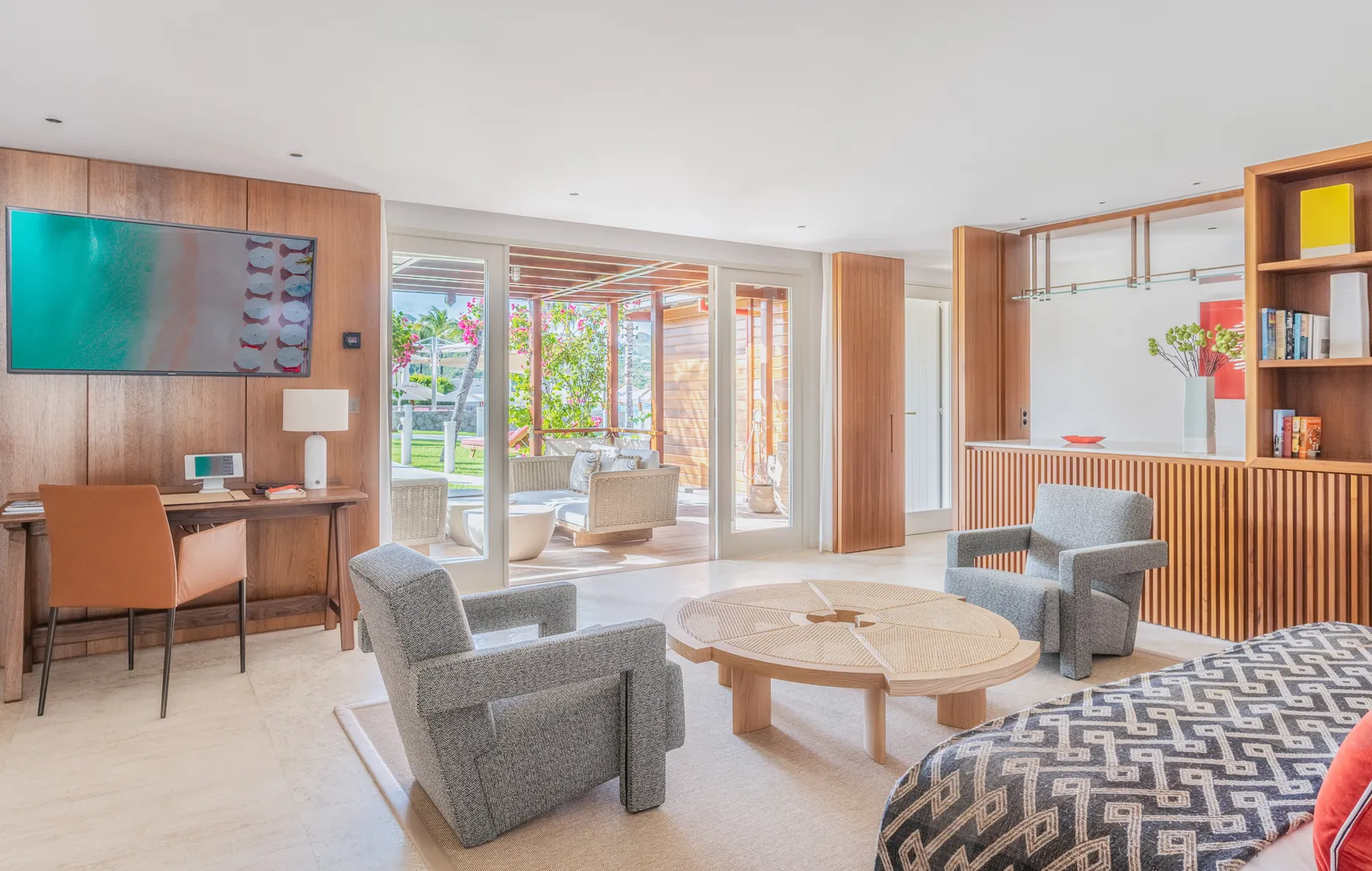 Beach House Frangipani’s living room: timber-panelled walls, 2 patterned grey armchairs, a round table and glass doors to the garden.