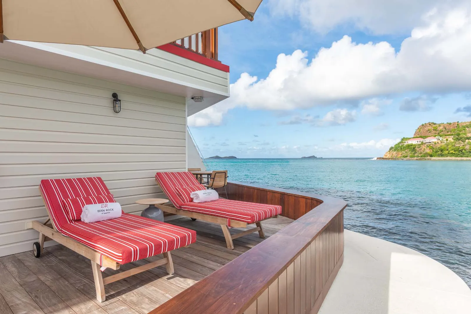 The Captains Room’s terrace: 2 red-striped loungers under a parasol, straight above the sea, with the headland opposite.