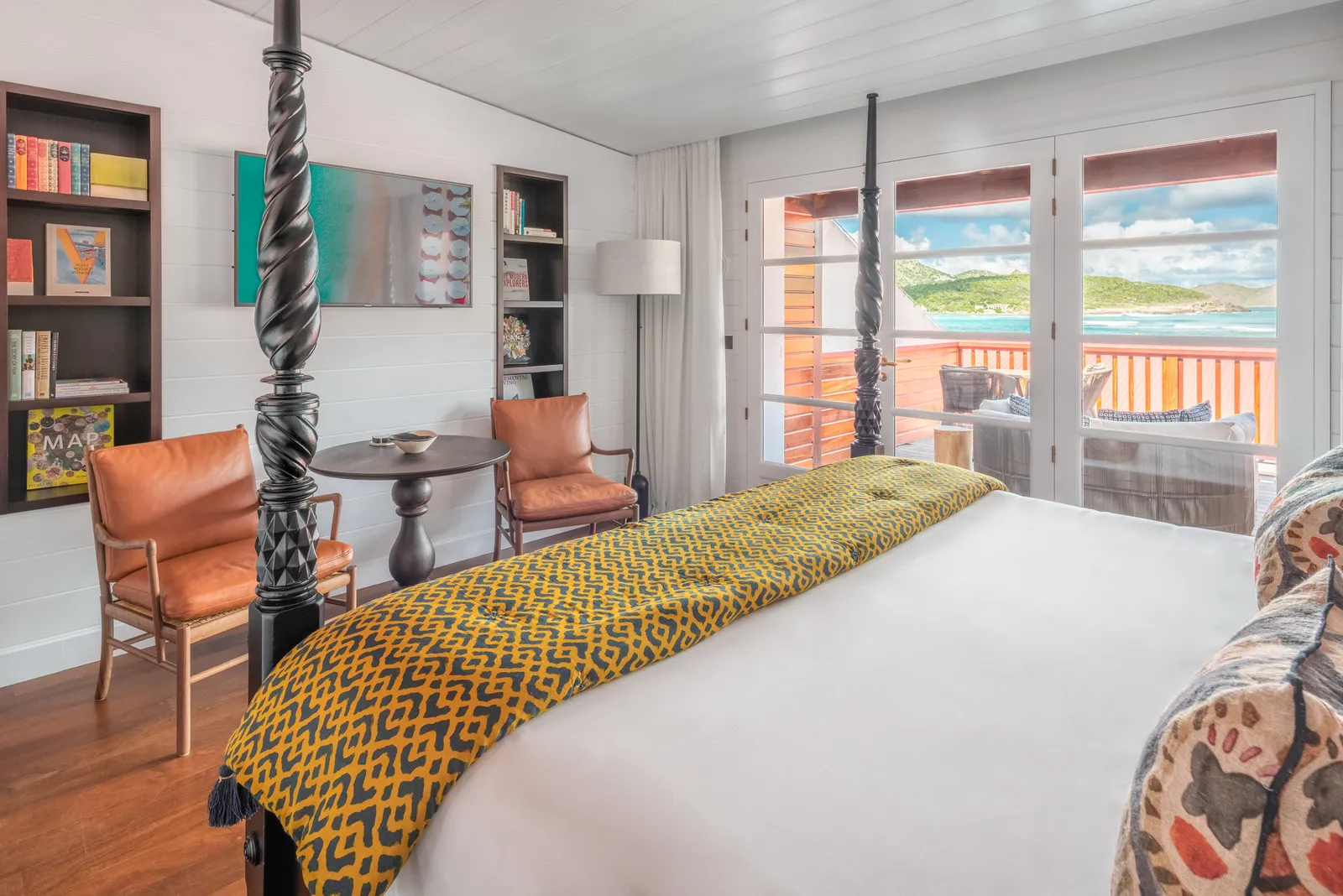 An Eden Room: the dark four-poster with a yellow throw, bookshelves, 2 leather armchairs and the door open to a terrace over the bay.