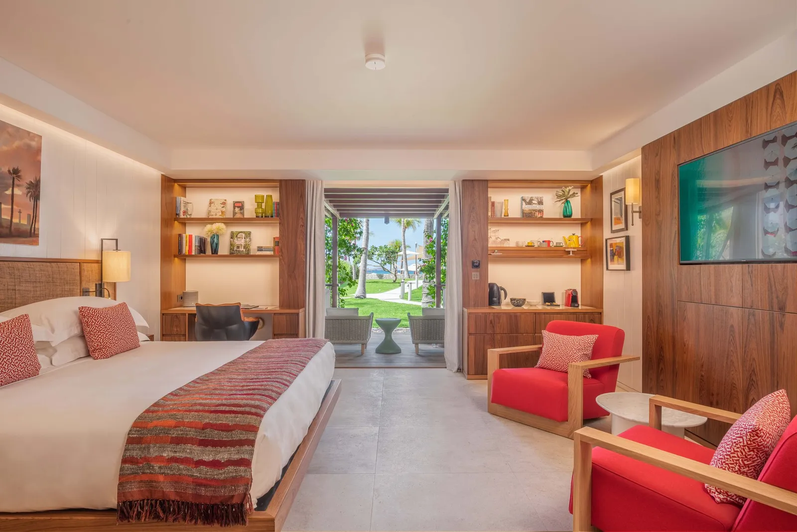 A Plantation Room: the bed, timber-panelled walls with shelves, 2 red armchairs and the door open to the palm garden.