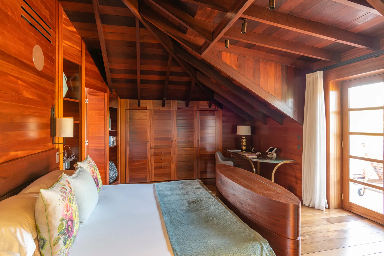 A bedroom of Signature Suite Howard Hughes under its dark timber pitched roof, with panelled walls and a door to the terrace.