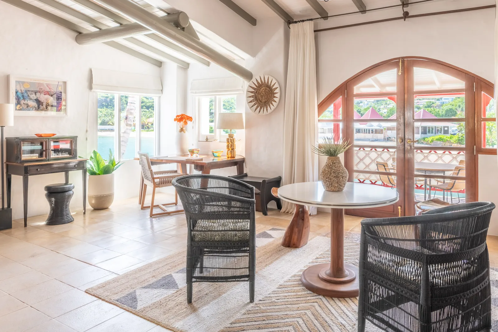 Spencers Room’s sitting area under white beams: a round table, black woven chairs and arched doors to the balcony and the bay.