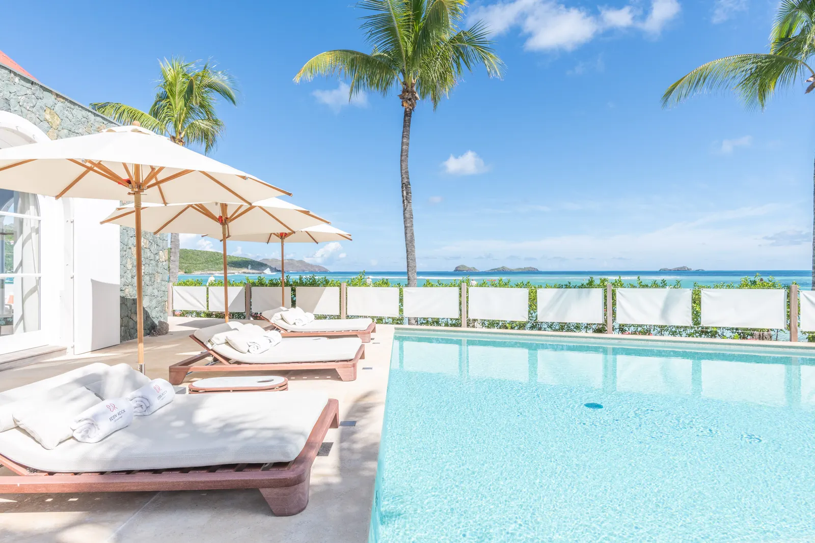 Villa Nina’s pool, with loungers, white parasols and palms, and the sea beyond the fence.